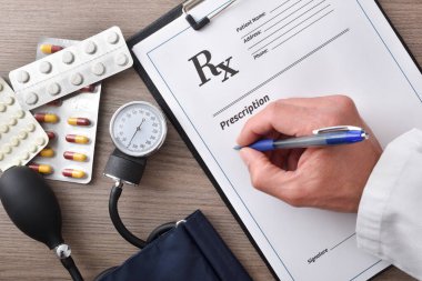Doktor, ahşap bir ofis masasında, tansiyon monitörü ve ilaçları olan bir belgeye kan basıncı için reçete yazıyor. Üst görünüm.
