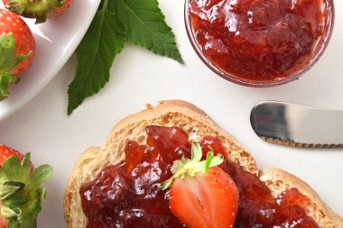 Çilek reçelinin ayrıntıları bir dilim ekmeğe ve beyaz bir masada üzerinde çilek ve bıçak bulunan cam bir konteynıra yayıldı. Üst görünüm.