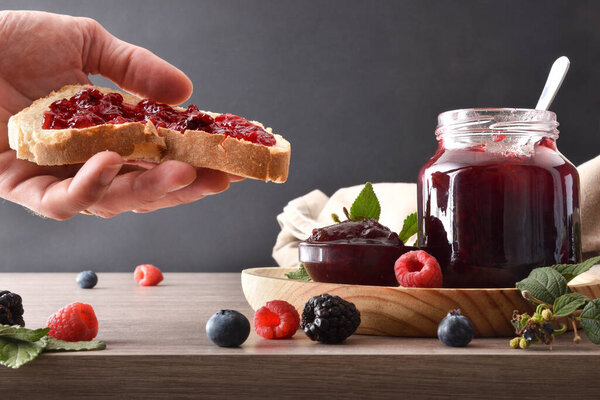 Hand showing slice of bread with berries jam and wooden kitchen bench with containers with jam and breakfast products and isolated dark background. Front view.