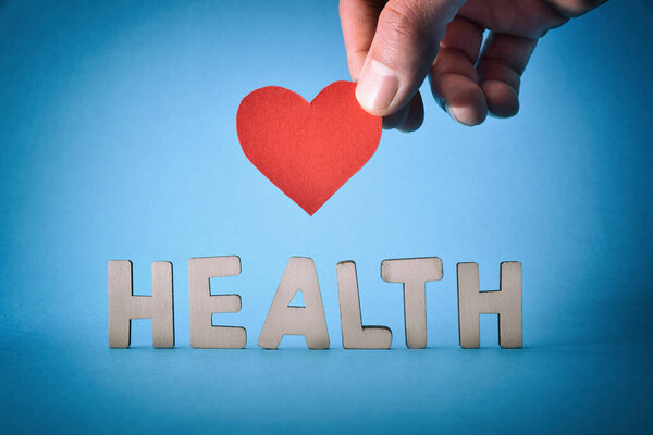 Рука держит красную бумагу сердце вырезывание и деревянные буквы со словом здоровье и синий градиентный фон.