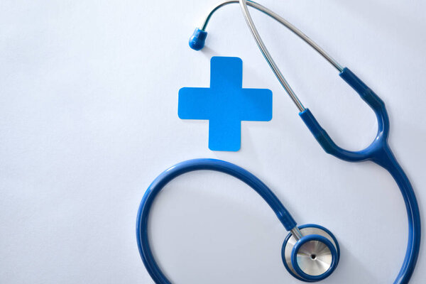 Health background with stethoscope and blue cross isolated on white table. Top view