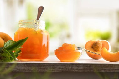 Kavanoz ve kase şeftali reçeli ve meyve parçaları ve arka planda ahşap masa ve mutfak yapraklı dilimler. Ön görünüm.