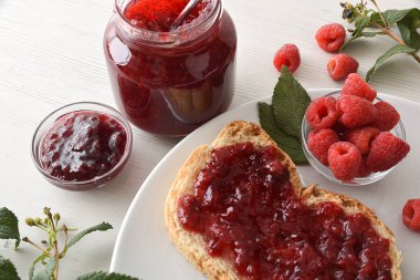 Beyaz tabakta ahududu reçeli, böğürtlen dolu bir tost ve ahşap bankta reçel kavanozu. Üst Görünüm.