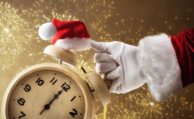 Santa's hand in a suit and glove holding a clock that shows 12 o'clock decorated with a Santa hat and golden fireworks in the background. clipart