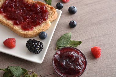 Kavanozlar ve böğürtlenlerle dolu ahşap bir bankta, üzerinde orman meyvesi reçeli tost. Üst Görünüm.