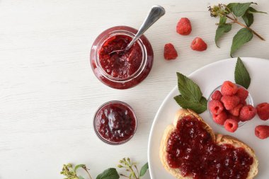 Ahududu reçeli kavanozlu beyaz ahşap mutfak tezgahı ve reçelli tost ve bir kase böğürtlen ile hazırlanmış beyaz tabak. Üst görünüm.