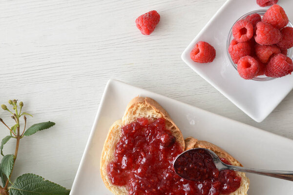 Тост с малиновым вареньем на белой тарелке свежеприготовленный на завтрак и миска, полная ягод на белой деревянной скамейке. Вид сверху.