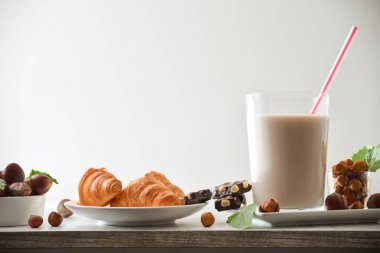 Ahşap mutfak tezgahında samanla birlikte bir bardak fındık içeceği, etrafında meyveler ve çikolatalar ve izole edilmiş arka planda kruvasanlar. Ön görünüm.