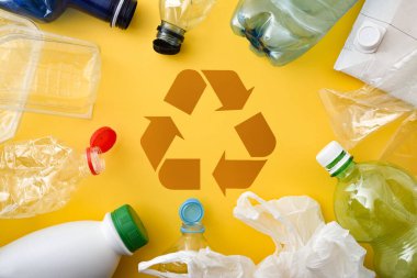 Plastik ve ambalaj kirliliği ve geri dönüşüm konsepti sarı bir taban, şişeler ve konteynırların merkezinde geri dönüşüm sembolü ile. Üst görünüm.