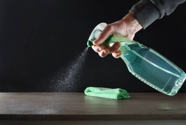 Ev temizliği için yeşil mikrofiber bez ve karalahana kaplı ahşap bir mobilyaya uygulanan bir ürünle el ele. Ön görünüm.