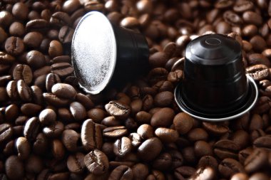 Kavrulmuş kahve çekirdeğinin üzerinde iki siyah alüminyum espresso kapsülü detayı. Üst Görünüm.