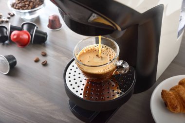 Kapsül kahve makinesi, kahve kapsülleri ve hamur işleriyle birlikte ahşap mutfak tezgahında cam bir fincanda espresso hazırlıyor. Yüksek görünüm.