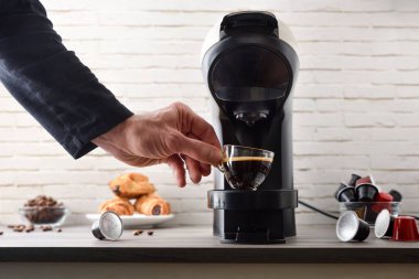 Kahve kapsülleri, fasulyeler, hamur işleri ve beyaz tuğla arka planlı ahşap bir bankta duran kapsül espresso makinesinden taze demlenmiş bir fincan kahve almak. Ön görünüm.