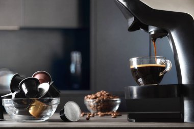 Kapsül kahve makinesi, arka planda kahve kapsülleri ve koyu mutfak olan ahşap mutfak tezgahında cam bir fincanda espresso hazırlıyor. Ön görünüm.
