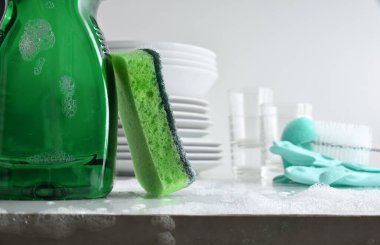 Bulaşık makinesinin dibinde, içinde ürün olan bir şişe ve aşındırıcı ve sünger yüzeyi olan bir temizlik yastığı beyaz mutfak tezgahında su, köpük, bulaşık ve temizlik malzemeleri olan bir tabaka..