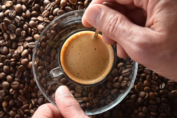 Cam bir fincanda taze demlenmiş kahve ve kahve çekirdeğinin üzerinde metal kaşıkla çay tabağı karıştıran ellerin ayrıntıları. Üst görünüm.