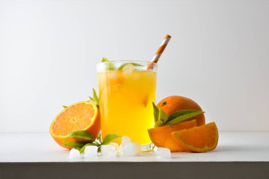 Pipetle süslenmiş cam bardakta buzla portakal suyu ve beyaz bir masada meyve, buz ve beyaz izole arkaplanlı yapraklarla bir dilim meyve. Ön görünüm.
