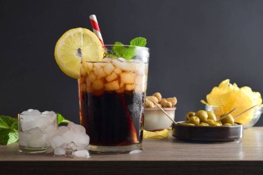 Nane yaprağı ve siyah arkaplanla izole edilmiş limon dilimleri ile süslenmiş fıstık ve zeytin gibi abur cuburlarla ahşap bir masada kola içmek. Ön görünüm.