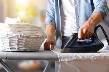 Beyaz bir gömleği ütü masasında ütüleyen zarif bir adamın ayrıntıları. Bir oturma odasında içi yeni ütülenmiş gömleklerle dolu bir sepetle. Ön görünüm.