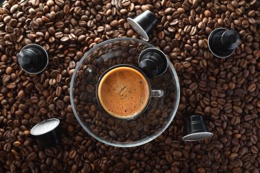 Ortadaki cam bir tabakta köpüklü kahve fincanı kızarmış kahve çekirdekleri ve siyah alüminyum kahve kapsüllerinin üzerinde. Üst görünüm.