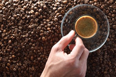 Hand holding a cup full of coffee to enjoy on a glass plate on top of a pile of coffee beans. Top view.
