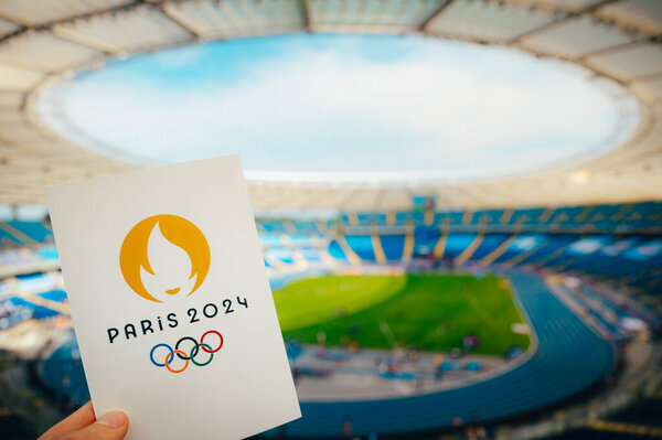 PARIS, FRANCE, JULY 7, 2023: Embracing the Symbol: Athlete Holds Iconic Emblem of Paris 2024 Summer Olympics, with Modern Stadium as a Majestic Backdrop. A Symbolic Prelude to Paris 2024 Olympics