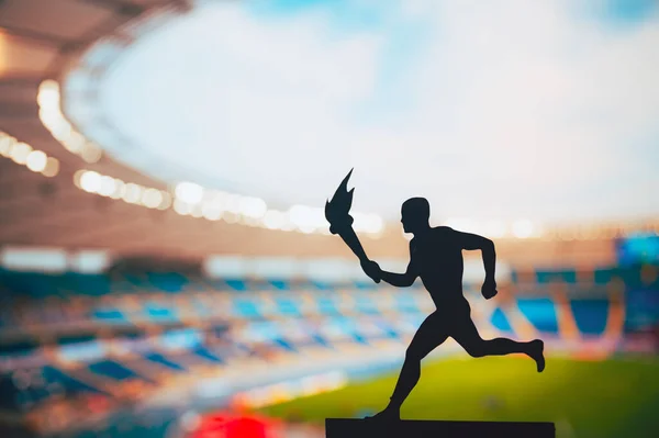 Igniting the Spirit: Silhouette of Male Athlete Carrying the Torch Relay. Modern Track and Field Stadium as the Striking Backdrop. Capturing the Essence of Summer Games 2024 in Paris