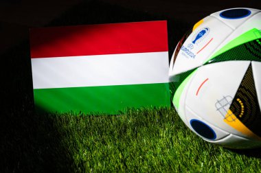 BERLIN, GERMANY, APRIL 17, 2024: Hungary national flag and official soccer ball of Euro 2024 football tournament in Germany placed on green grass. Black background, edit space clipart