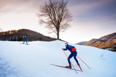 Düzenleme alanı olan bir kış sporu fotoğrafı. Dusk 'taki İskandinav Kayak Patikasını Kucaklayan İki Dişi Sporcu Kış Günbatımının Çerçevelediği