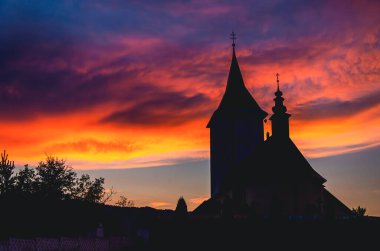 Çan kulesi ve üzerinde haç olan bir kilise. Gökyüzü turuncu ve güneş batıyor.