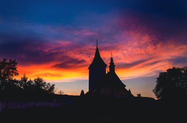 Üzerinde uzun bir çan kulesi ve haçı olan bir kilise. Gökyüzü turuncu ve güneş batıyor.
