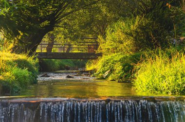 Üzerinde köprü olan küçük bir dere. Köprü ahşap ve birkaç bank var. Su berrak ve çimenler yeşil.