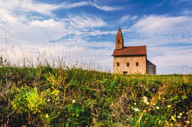 Çan kulesi olan küçük kırmızı bir bina yamaçta duruyor. Bina çimenler ve çiçeklerle çevrili.