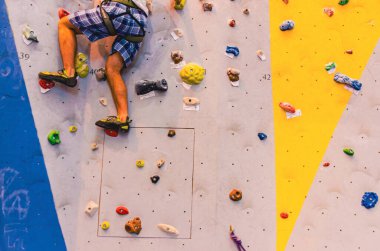 Bir adam kaya duvarına tırmanıyor. Duvar birçok farklı renkli kayalardan yapılmıştır. Adam sarı bir gömlek giyiyor.