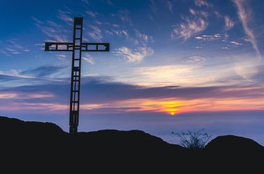 Bir haç tepenin yamacında duruyor ve arkasında güzel bir günbatımı var. Gökyüzü bulutlarla dolu ve güneş uzaklardan batıyor.