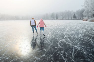 A couple dressed in soft pastel colors skate together on a frozen pond, embraced by a misty winter landscape and the warm magical sun, celebrating the Christmas season