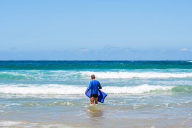 Genç adam denize sörf tahtasıyla giriyor, su sporu yapıyor, güneş gömleği giyiyor, deniz manzarası seyrediyor, yaz güneşli bir gün..