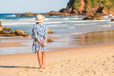 Şapkalı, mavi ve beyaz elbiseli genç bir kadın kumlu bir sahilde yürür, güneşli bir yaz gününde deniz manzarası.