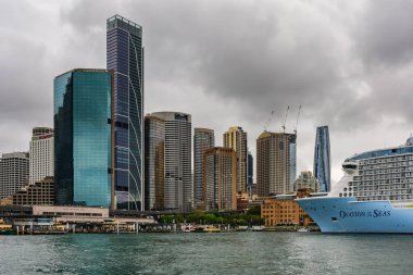 Sydney, Avustralya - 03 Mart 2023: Circular Quay, kıyıya demirlemiş yolcu gemisi, şehir merkezinde cam gökdelenler, nehir üzerindeki feribottan manzara.