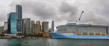 Sydney, Avustralya - 03 Mart 2023: Circular Quay, kıyıya demirlemiş yolcu gemisi, şehir merkezinde cam gökdelenler, nehir üzerindeki feribottan manzara.