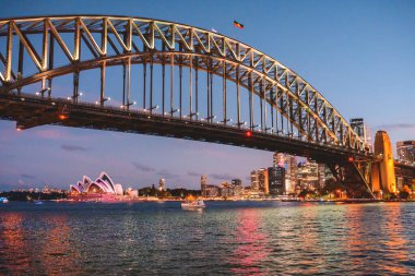 Sydney, Avustralya - 04 Mart 2023: Sydney Liman Köprüsü ve Sydney Opera Binası gece renkli ışıkla aydınlatılmış, nehirdeki bir teknenin manzarası.