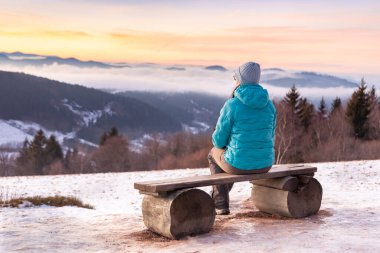 Genç bir kadın ahşap bankta oturuyor ve dağlarda renkli günbatımını seyrediyor. Vadide yoğun sisli kış dağları manzarası..