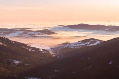 Doğu Sudetleri, dağlarda kış yürüyüşü sırasında tepenin zirvesinden vadideki yoğun sisli dağ manzarası, turuncu günbatımında dağlardaki geniş kış manzarası.