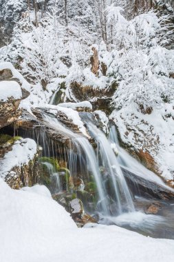 Dağ nehrinde küçük bir şelale, karla kaplı kayalardan aşağı su akıyor. Kış manzarası bir dağ yürüyüşü patikasında.