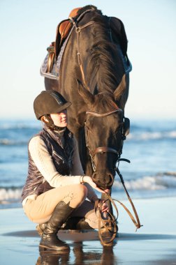 Genç bayan binici deniz / okyanus kıyısındaki güneş ışınlarında kestane atının yanında çömelmiş oturuyordu. Çok renkli açık havada dikey yaz görüntüsü