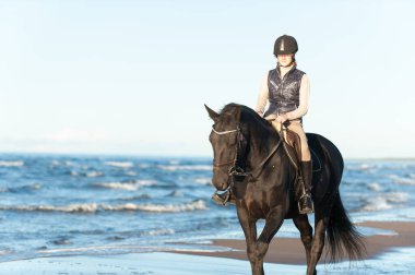 Genç bayan binici, deniz / okyanus kıyısında güneş ışığında ata biniyor. Çok renkli, açık havada yatay yaz manzarası ve arka plan ışığı.