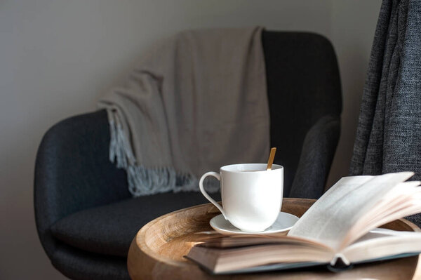 Cozy textile armchair with warm blanket and opened book with ceramic cup of tea on wooden coffee table for relaxing evening. Lifestyle indoor horizontal colored image.