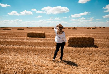 Hasattan sonra ekili buğday tarlasında yürüyen kadın çiftçi. Altın tarlalarda buğday yığını ve mavi gökyüzü olan güzel sarışın kadın. Boşluğu kopyala