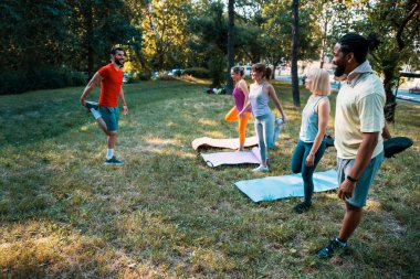 Yoga yapan çok ırklı arkadaşlar. Antrenörlü insanlar parkta yeşil çimlerde antrenman yapıyorlar. Beden eğitimi sınıfındaki çeşitli insanlar dışarıda egzersiz yapıyor. Yaz günü birlikte idman yaparız. Boşluğu kopyala