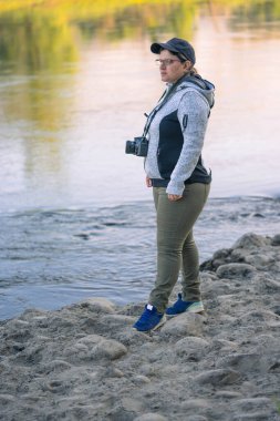 Sahilde duran bir kadın kamerayla güneşin dokunduğu inanılmaz dağlara bakıyor.